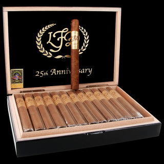 An open cigar humidor box with ten wrapped cigars inside and one uncapped cigar standing upright on the black lid. The lid features a black background with a gold wreath and the text '25th Anniversary' and 'L.